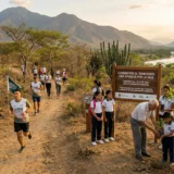 Fotografía de la carrera atlética "Correr por el Territorio" en el ecosistema de bosque seco tropical del Valle del Patía. En un sendero de tierra, varias personas corren mientras que, a la derecha, un grupo de niños y adultos siembran un árbol junto a un letrero de madera del evento. Al fondo se observa el cauce del río Patía y un paisaje montañoso.