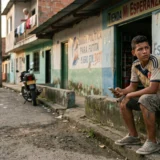 Un joven con una camiseta desgastada de la selección de fútbol de Colombia se sienta en un andén de concreto en una calle de barrio humilde, sosteniendo un teléfono móvil y mirando atentamente hacia un lado con expresión alerta. Al fondo, una tienda local con la fachada despintada dice "Tienda Mi Esperanza" y tiene grafitis. Más allá, un grupo de jóvenes está de pie en la calle y una persona mayor observa desde un balcón. La iluminación es natural de atardecer.