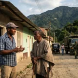 Dos hombres conversando en una calle de tierra de un pueblo de montaña. Un hombre mayor lleva un poncho tradicional y sostiene un bastón, mientras que un hombre más joven con una gorra gesticula. Al fondo, una patrulla de soldados armados camina por la calle, con un camión militar y mototaxis estacionados cerca.