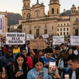 Fotografía documental de una multitud densa y alienada en la Plaza de Bolívar, Colombia, al atardecer. Personas diversas miran teléfonos inteligentes que brillan, ignorando su entorno y a los demás. Están rodeadas de carteles electorales simplistas como 'SOLUCIONES SIMPLES A PROBLEMAS COMPLEJOS' y 'MANO DURA, LA NUEVA DERECHA' y propaganda de 'DE LA ESPRIELLA 2026'. Muestra abdicación de pensamiento autónomo por inmediatez digital.