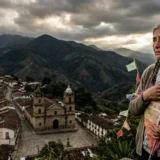 Una mujer de edad avanzada con trenzas, vestida con una chaqueta de lana, llora en un mirador de montaña. Sostiene una cometa de papel de colores con el texto '9 DE ABRIL - MEMORIA' escrito a mano. Al fondo, un pueblo tradicional colombiano con una iglesia y montañas dramáticas bajo un cielo nublado