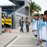 Entrenador argentino consuela a un jugador juvenil cabizbajo en la zona de buses de un estadio moderno, mientras al fondo se observa la celebración espontánea de los jugadores de la selección colombiana.