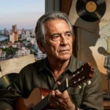 Hombre de cabello canoso sostiene una guitarra junto a discos de vinilo y un paisaje costero.