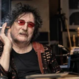 Charly García con gafas rojas sentado junto a un piano en un estudio musical