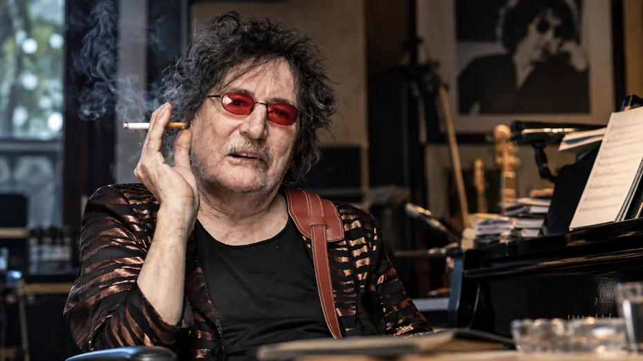 Charly García con gafas rojas sentado junto a un piano en un estudio musical