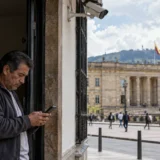 Hombre utiliza un telefono movil cerca de una camara de vigilancia frente a un edificio institucional