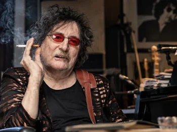 Charly García con gafas rojas sentado junto a un piano en un estudio musical